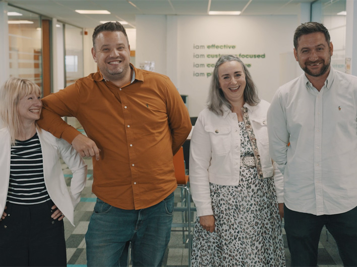 Four cheerful employees at iamproperty, three standing and one leaning in, with positive affirmations like 'I am effective' and 'I am customer-focused' visible in the background.