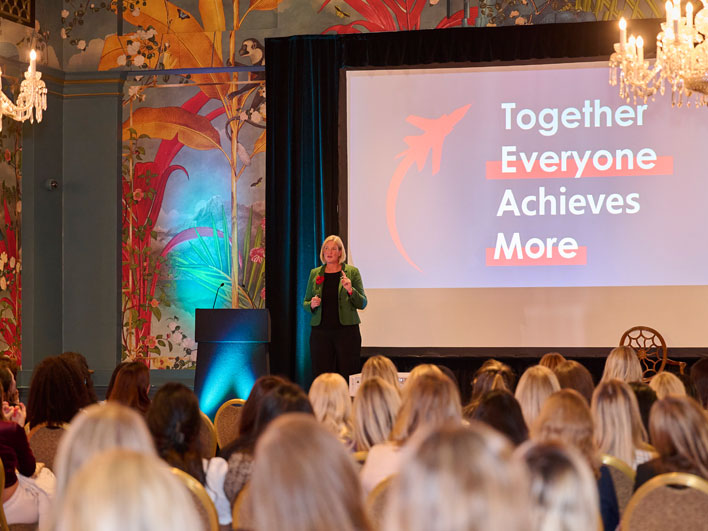 A woman presenting at a conference with the projection 'Together Everyone Achieves More' on the screen behind her, in a room with an audience and ornate, colorful wallpaper.