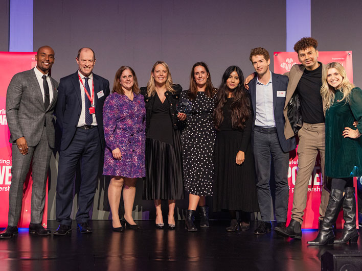 A diverse group of eight award recipients standing onstage, smiling with a trophy, at the PT Award event, with event banners in the background.