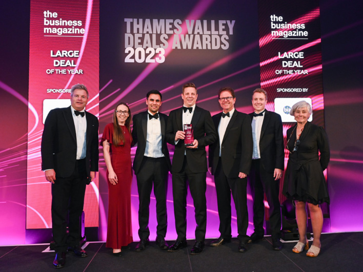 A team of professionals, dressed in formal attire, proudly holding a trophy at the Thames Valley Deals Awards 2023 event, with event branding visible in the background.