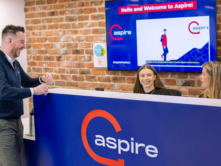 Two employees at the Aspire reception desk smiling and interacting with a colleague, with a welcoming digital display screen in the background and a brick wall interior.
